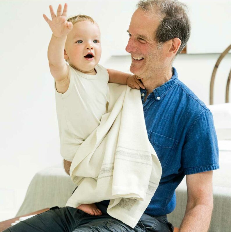 A proud grandpa with baby holding Swans Island's Grace baby blanket, handwoven in undyed ivory soft organic merino wool with classic undyed grey wool stripes and a soft silk border.