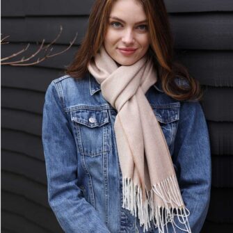 Swans Island Company's Baby Alpaca Scarf is woven in Peru with the softest baby alpaca. Lightweight and generously proportioned, this scarf has a classic herringbone twill pattern with fringed edges. . Shown here in Camel with natural, on a female model wearing a jean jacket.